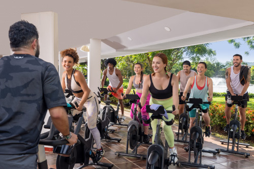 Grupo de personas que están sobre bicicletas de spinning se ejercitan en el gimnasio, al cuál se puede acceder a través de las tarifas y planes en el hotel Lagosol