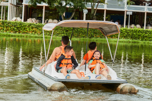 Dos adultos y dos niños están navegando en un bote de color blanco, en un lago que está rodeado de naturaleza en las instalaciones del hotel y parque acuático Lagosol