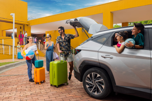 Familia de cinco personas baja las maletas de diferentes colores de un carro de color gris, frente a las instalaciones color mostaza de Compensar, en donde se realizo una reserva de habitación en el hotel Lagosol