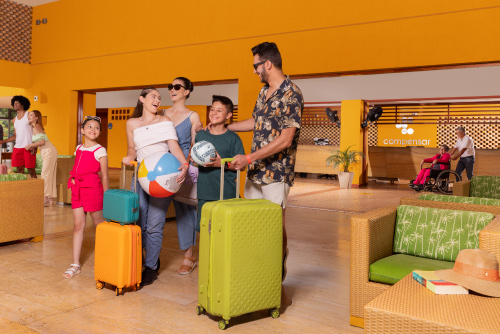 Familia entrando con maletas a un lobby que tiene paredes de color mostaza y sillones con cojines de color verde en donde hacen el pago de su reserva de habitación en el hotel Lagosol