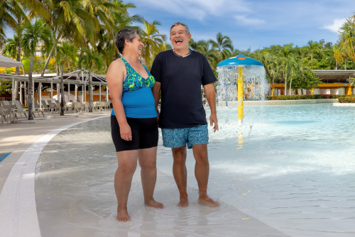 Pareja de adultos mayores, que usa ropa cómoda, va caminando junto a una piscina de agua cristalina que está rodeada de gran cantidad de vegetación y hace parte de los planes turísticos de Lagosol