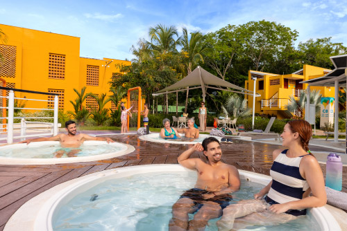 Pareja está disfrutando dentro un jacuzzi que hace parte de la oferta de planes en el hotel Lagosol, ubicado al aire libre, rodeado de árboles y edificios de dos pisos de color mostaza