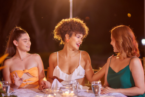 Tres mujeres que usan vestidos de diferentes colores están cenando en un lugar elegante en medio de la noche, luego de adquirir un paquete con la mejor variedad de tarifas y planes del hotel Lagosol Compensar
