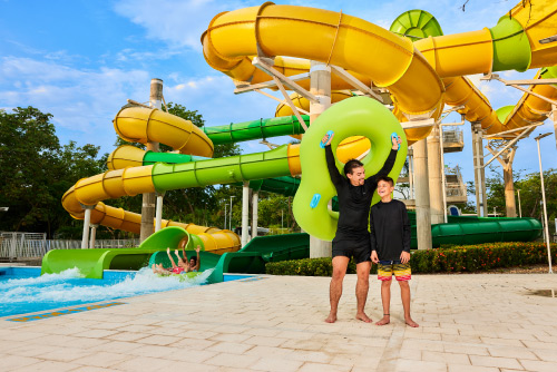 Un hombre y un niño están frente a un gran tobogán de color amarillo y verde que está rodeado de gran cantidad de naturaleza, en las instalaciones del hotel Lagosol Compensar
