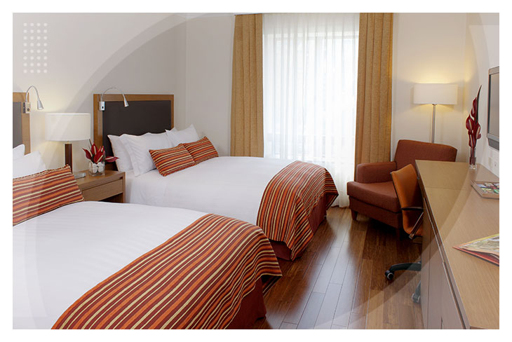 Dos hombres y dos mujeres, que visten ropas de colores vinotinto, café, azul oscuro, azul claro, negro, blanco y amarillo mostaza, están recostados en una cama blanca de una de las habitaciones del Hotel Lagomar, junto a ellos hay dos mesas de noche. Uno de los hombres observa y sostiene una tablet, y una de las mujeres sujeta un control de televisión