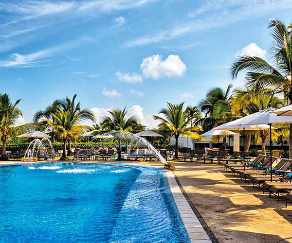 Piscina de agua cristalina, con tres chorros de agua, rodeada de gran cantidad de sillas asoleadoras cafés, cada una con su parasol de color blanco, forma parte de la oferta de hoteles Estelar en Colombia.  Alrededor gran cantidad de palmeras de tamaño medio de color verde con amarillo en un día soleado y con cielo azul