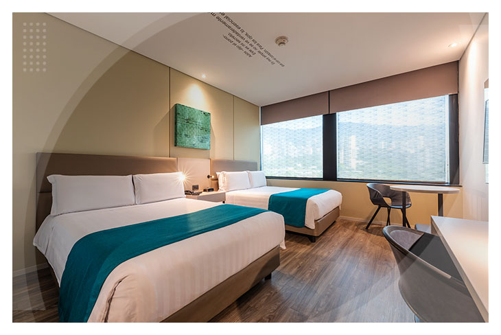 Dos hombres y dos mujeres, que visten ropas de colores vinotinto, café, azul oscuro, azul claro, negro, blanco y amarillo mostaza, están recostados en una cama blanca de una de las habitaciones del Hotel Lagomar, junto a ellos hay dos mesas de noche. Uno de los hombres observa y sostiene una tablet, y una de las mujeres sujeta un control de televisión
