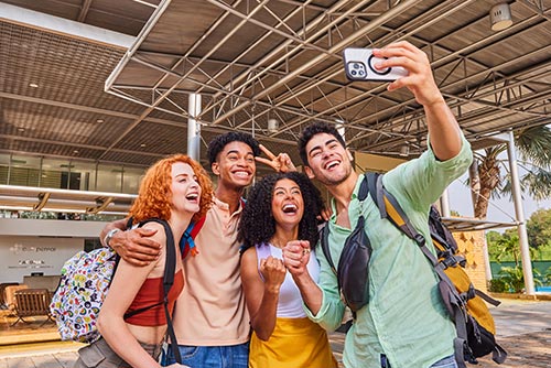 Dos mujeres y dos hombres, que utilizan morrales y ropa para clima cálido de colores blanco, durazno, amarillo, rojo, verde claro, azul y gris, están de pie sonriendo para tomarse una selfie frente a la recepción del Hotel Lagomar