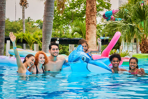 Tres mujeres y dos hombres, que utilizan trajes de baño de color magenta, azul y negro, se encuentran sonriendo dentro de una piscina de Lagomar alrededor de un niño que está sobre un flotador con forma de tiburón azul y blanco