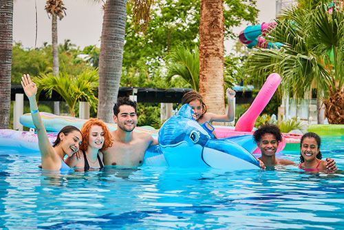 Familia de seis personas sonriendo dentro de una piscina mientras juegan con un flotador de ballena y disfrutan de uno de los planes en oferta para ir a Lagomar y Lagosol. Alrededor hay palmeras, árboles y se alcanza a ver una parte de un tobogán morado y azul