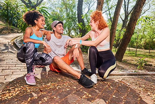 Un hombre y dos mujeres de cabello rizado, que visten ropa deportiva de colores negro, blanco, naranja y cian, están sentados al aire libre en un camino del Hotel Lagomar rodeados por árboles