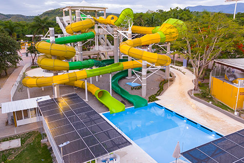 Gran tobogán amarillo con verde que desemboca en piscina, la cual está rodeada con techos de vidrio, debajo de los cuales hay sillas asoleadoras que hacen parte de la oferta en el hotel Lagosol 6 días. Alrededor hay mucha naturaleza