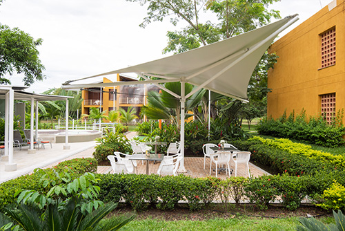 Foto de las instalaciones de uno de los hoteles Compensar, en la que se observan zonas verdes, sillas blancas, quioscos y edificios de color mostaza