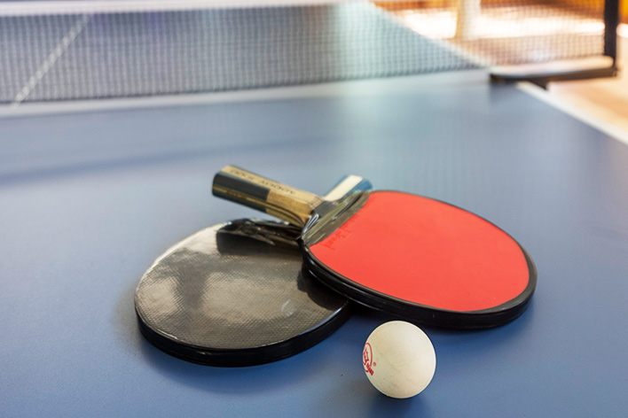 Sobre una mesa de ping pong azul, hay una pelota blanca y dos raquetas, una negra y la otra roja, que pertenecen a los planes vacacionales en Lagosol