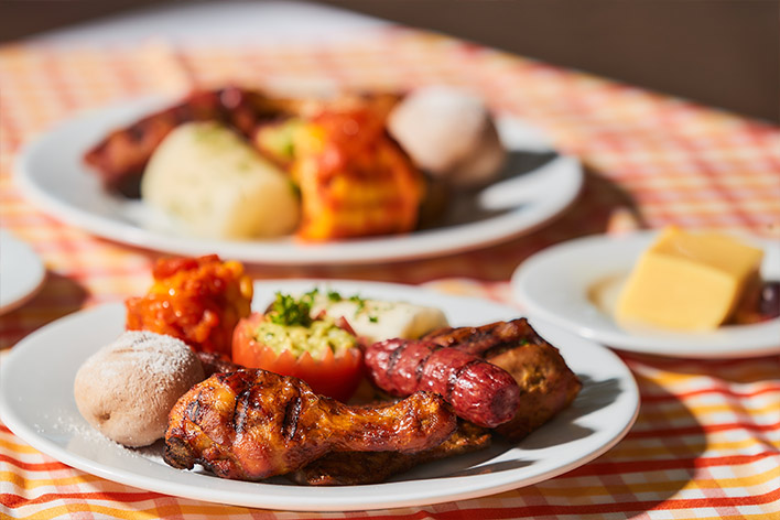 Fotografía de tres platos blancos con una presa de pollo asado, papa salada, un tomate y otros alimentos que ofrece el hotel y parque acuático Lagosol. Los platos están sobre un mantel de cuadros rojos con naranja