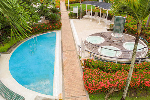 Toma elevada de una pequeña piscina, desde la cual se alcanzan a ver tres jacuzzis, una ducha, cuatro sillas asoleadoras, a las cuales se puede acceder con el plan living Lagomar. Alrededor se ven flores rojas, palmeras y vegetación