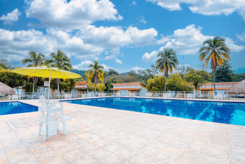 Dos piscinas, una grande y una mediana, de las instalaciones del hotel y centro vacacional Las Palmeras con una silla de vigilancia con sombrilla amarilla, árboles, palmeras grandes, y casetas alrededor del lugar