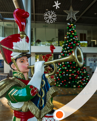 En una recepción hay un muñeco navideño tocando una trompeta, también, hay un árbol de Navidad  y personas caminando por el lugar