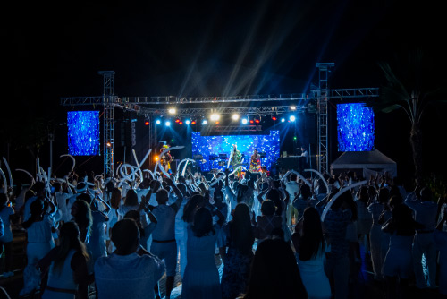Personas, vestidas de blanco, con globos en la mano, disfrutando de un evento musical, donde hay luces y visuales azules