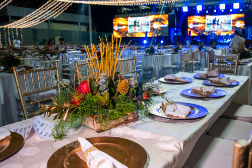 Mesa elegante decorada con centros de mesa navideños, platos dorados y servilletas blancas, como parte del montaje para una cena de Navidad en un evento nocturno