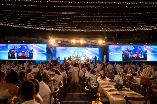 Gran evento social con asistentes vestidos de blanco, sentados en mesas organizadas frente a pantallas gigantes y decoraciones con luces colgantes, durante una cena nocturna