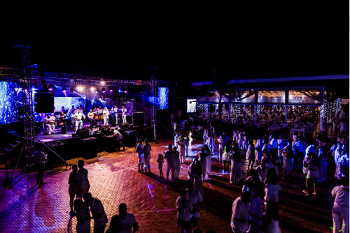 Personas, disfrutando de un evento musical en vivo, están en un lugar con tarima en el que se observan visuales azules en las pantallas
