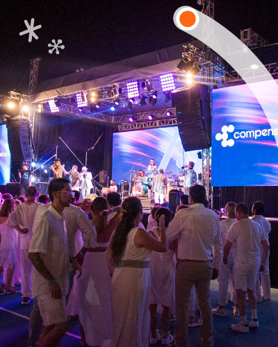 Personas, vestidas de blanco, están bailando y escuchando un concierto de una agrupación musical, con visuales azules y el logo de Compensar