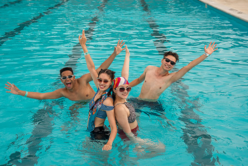 Cuatro personas, dos hombres y dos mujeres dentro de una piscina levantando los brazos con intención de celebración mientras disfrutan de las ofertas para afiliados Compensar en Lagosol y Lagomar