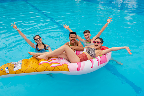 Dos hombres y dos mujeres, compartiendo dentro de una piscina, una de ellas sobre un flotador en forma de helado, levantan los brazos para celebrar, mientras disfrutan de los planes en oferta para ir a Lagomar y Lagosol