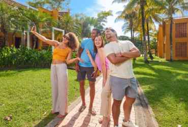 Cuatro personas, tomándose una fotografía en medio de un sendero que está rodeado de naturaleza y edificios de color mostaza, los cuales hacen parte de la oferta de  hospedaje en el hotel Lagosol. En el fondo se ven algunas nubes y un cielo azul soleado