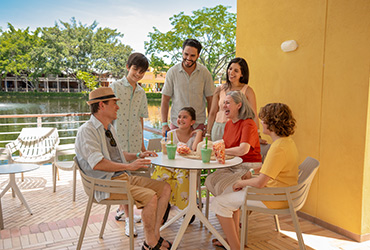 Fotografía de siete personas en la zona de descanso de su hospedaje en hotel Lagosol de Compensar, en donde cuatro están sentadas alrededor de una mesa, tomando jugo y comiendo rosquitas de queso, y detrás de ellos hay otras tres personas de pie sonriendo