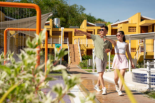 Un hombre y una mujer, que visten ropa de colores verde oliva, gris, blanco y rosado claro, están caminando y señalando una fuente de agua naranja en Lagosol; detrás de ellos hay un edificio amarillo y vegetación