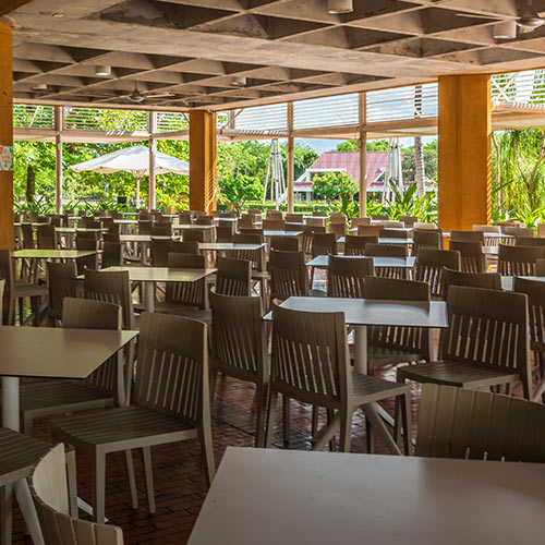 Salón lleno de mesas con sillas y grandes ventanales en el comedor donde se puede disfrutar de los beneficios del plan fin de semana festivo en Lagosol. En el exterior se puede ver un parasol blanco, un techo vinotinto y gran cantidad de árboles