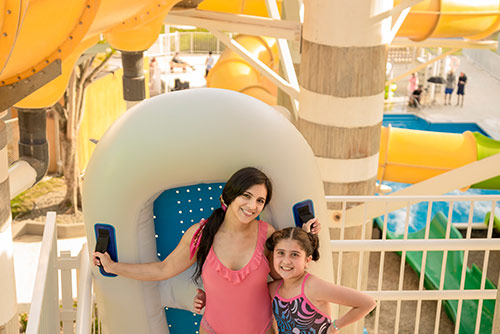 Una mujer y una niña, que usan vestidos de baño de color rosado y estampado morado con azul, están de pie abrazándose y sosteniendo un flotador doble blanco con azul; detrás de ellas hay una valla blanca y un tobogán amarillo