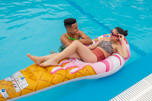 Una mujer, usando un vestido de baño, se encuentra sobre un flotador de piscina junto a un hombre con quien habla de los descuentos del fin de semana del Hotel Lagosol