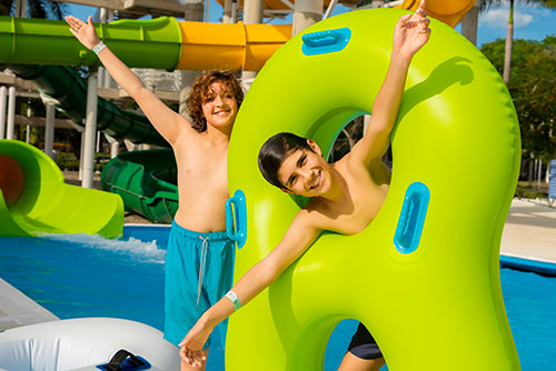 Dos adolescentes, usando traje de baño, están jugando con un flotador de color verde manzana al lado de una piscina con toboganes verdes y amarillos, mientras disfrutan de las reservas de temporada baja en Lagosol
