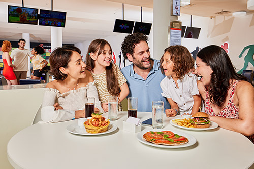 Dos mujeres, un hombre, un niño y una niña, que visten ropa de colores blanco, café claro, azul claro, rosado y rojo, están sentados frente a una mesa redonda blanca de la bolera del Hotel Lagomar, en la cual hay cuatro vasos con agua y gaseosa, tres platos blancos con una hamburguesa, pizza napolitana y una canasta de plátanos con salchichas