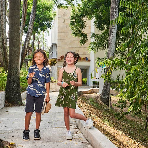 Un niño, que viste una camiseta, pantaloneta azul oscura y tenis negros, sostiene un sombrero tejido con su mano izquierda, mientras da un paseo por Lagomar junto a una niña, que usa un vestido verde musgo, con estampado de flores y tenis blancos, a su alrededor hay diferentes árboles