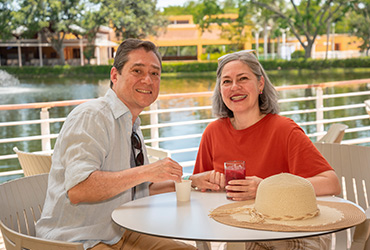 Hombre y mujer mayores están sentados en la zona de descanso frente a un lago, tomando jugo de mora y café en el hotel y parque acuático Lagosol; la mujer viste una blusa roja y el hombre una camisa gris