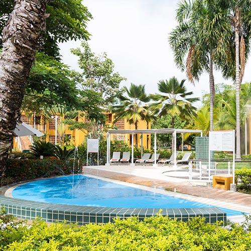 Pequeña piscina, rodeada de plantas, palmeras y seis sillas blancas para tomar el sol que hacen parte de la promoción de planes para ir a Lagomar. En el fondo se ve un edificio amarillo de dos pisos, en donde quedan las habitaciones del hotel