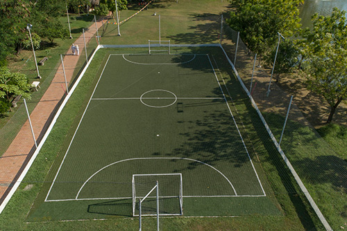 Cancha sintética de color verde, con claras demarcaciones de color blanco que hace parte de los descuentos en Lagosol y Lagomar. En el entorno hay un carril para caminar o trotar, postes de iluminación, sillas para descansar y el lugar está rodeado de árboles y vegetación
