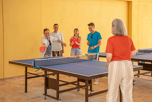 Cinco personas jugando ping pong en un salón de juegos incluido en las promociones de vacaciones Lagomar fin de semana. El lugar tiene paredes de color mostaza y hay dos mesas azules para jugar