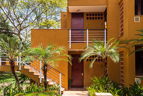 Edificio color mostaza de dos pisos, que tiene escaleras de rejas color blanco, en donde se puede comprar la promoción de planes para ir a Lagomar. Alrededor se pueden ver palmeras y árboles en un ambiente cálido