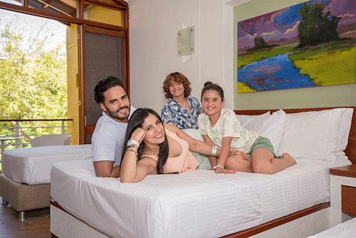 Familia de cuatro personas descansando en una cama con sabanas blancas la cual hace parte de los descuentos en hoteles Compensar por compras anticipadas. La habitación tiene paredes de color blanco, un cuadro con agua, árboles pintados. Hay un gran ventanal abierto desde el cual se alcanza a ver el exterior lleno de vegetación