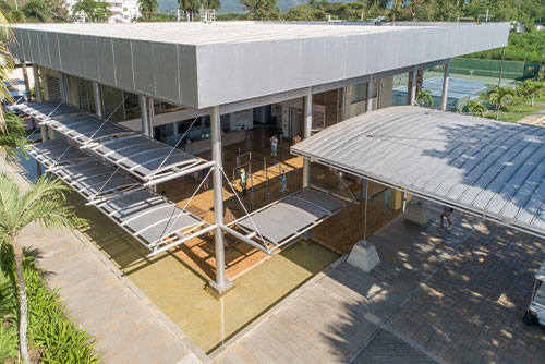Recepción del hotel con una gran estructura metálica gris, el lugar ideal para conocer los mejores planes en Lagomar con descuento. En el fondo, se puede ver una cancha de tenis de piso azul; todo esto rodeado de naturaleza en un día soleado
