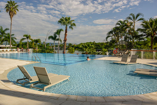 Amplia piscina con efecto infinito con una persona dentro que disfruta de la vista y de la promoción de reservas anticipadas de Compensar. En la piscina hay seis sillas asoleadoras adentro y en el entorno se pueden ver  palmeras y un lago