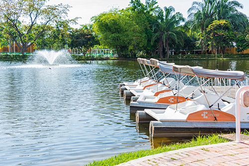 Amplio lago en el cual se pueden ver tres botes de pedales, cada uno con un parasol gris para proteger a las personas del sol, que se pueden usar luego de acceder a las promociones de vacaciones Lagomar fin de semana. Al fondo hay una fuente y gran cantidad de árboles que rodean el lago