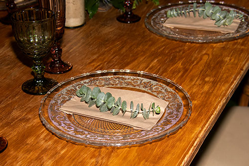 Mesa de madera con dos platos de vidrio tallados, cada uno con una servilleta y una planta sobre él, acompañados por una copa verde y una naranja, ambas vacías y talladas. Estos espacios se pueden disfrutar con los planes con descuentos en Lagosol y Lagomar. Sobre la mesa se alcanzan a ver otros recipientes y al lado el cojín de la silla