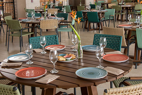 Comedor que tiene mesas de madera decoradas con platos de colores rojos y azules, copas de vino, flores y pan. Las sillas son artesanales y tienen espaldares verdes, cafés y azules, todo esto en un ambiente tranquilo y agradable, en donde podrás disfrutar tus comidas adquiriendo los planes en Lagomar con descuento