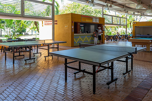 Mesas de ping pong debajo de una gran estructura con techo blanco dentro de las instalaciones del hotel en donde puedes disfrutar de los planes en Lagomar con descuento. Alrededor hay mesas para consumir alimentos, una mesa de billar y dos personas en la recepción